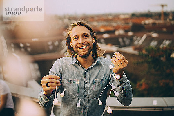 Porträt eines fröhlichen Mannes  der eine Leuchtschnur hält  während er auf einer Terrasse in der Stadt steht
