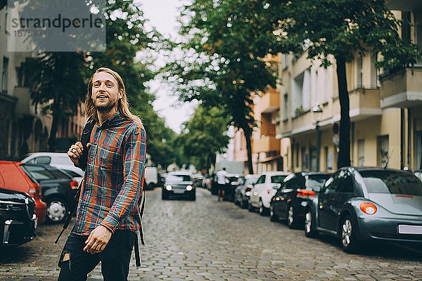 Junger Mann mit Rucksack beim Spaziergang auf gepflasterter Straße inmitten von Autos in der Stadt