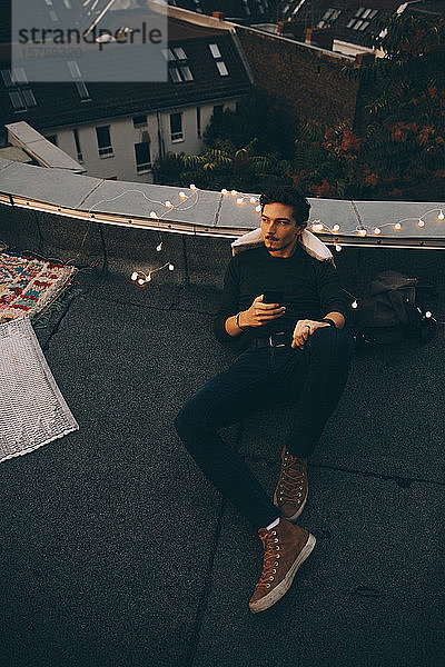 Hochwinkel in voller Länge eines nachdenklichen Mannes  der ein Mobiltelefon benutzt  während er sich während einer Party in der Abenddämmerung auf der Terrasse entspannt