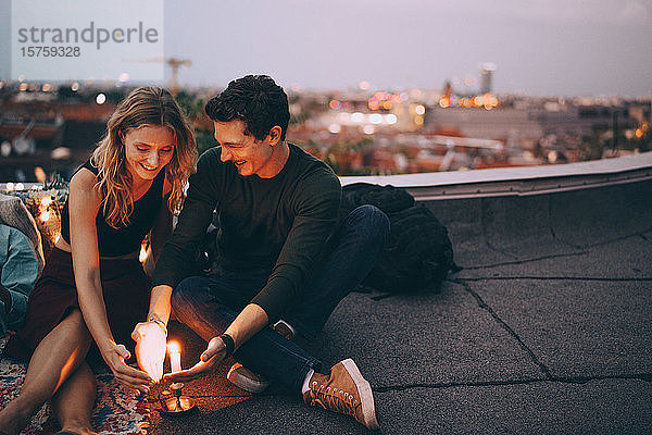 Lächelndes junges Paar schützt brennende Kerze mit den Händen  während es in der Dämmerung auf der Terrasse gegen den Himmel sitzt