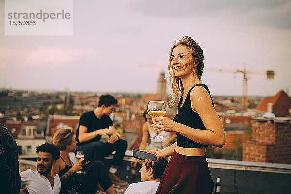 Lächelnde Frau genießt Getränk während einer Party mit Freunden auf dem Dach