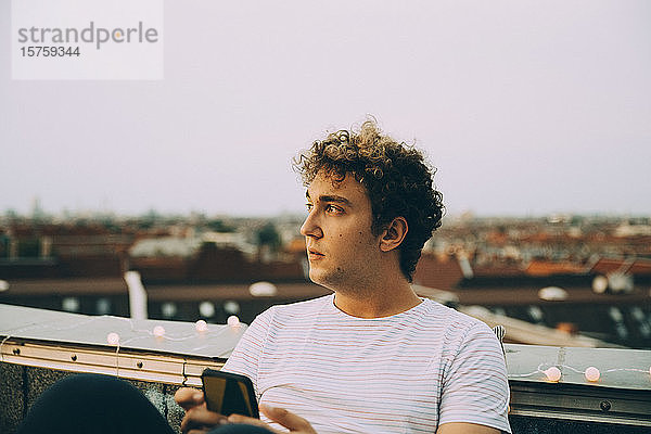 Nachdenklicher Mann schaut weg  während er sein Mobiltelefon auf einer Terrasse in der Stadt gegen den Himmel hält