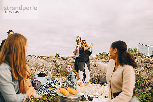 Glückliche männliche und weibliche Freunde  die sich beim Picknick auf dem Feld gegen den Himmel vergnügen