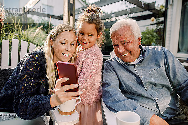 Lächelnde Großeltern und Enkelin beim Selbermachen mit dem Handy  während sie im Hinterhof sitzen