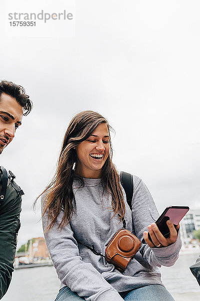 Lächelnde junge Frau benutzt Mobiltelefon  während sie mit einem Freund in der Stadt gegen den Himmel sitzt