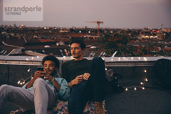 Nachdenklicher Mann sitzt mit einem Freund zusammen und benutzt ein Mobiltelefon auf einer Terrasse in der Stadt