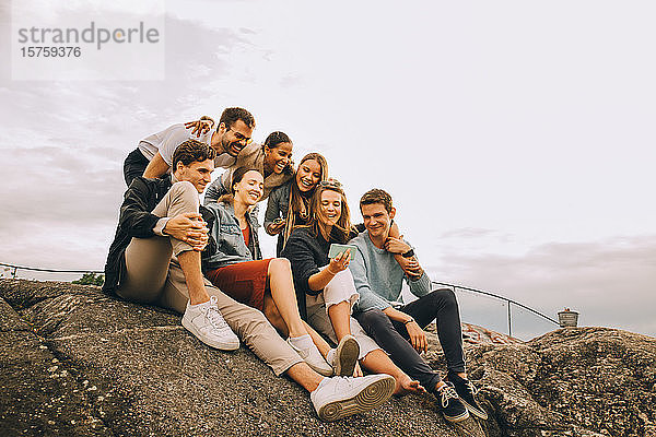 Glückliche männliche und weibliche Freunde schauen auf Mobiltelefone  während sie auf einer Felsformation gegen den Himmel sitzen