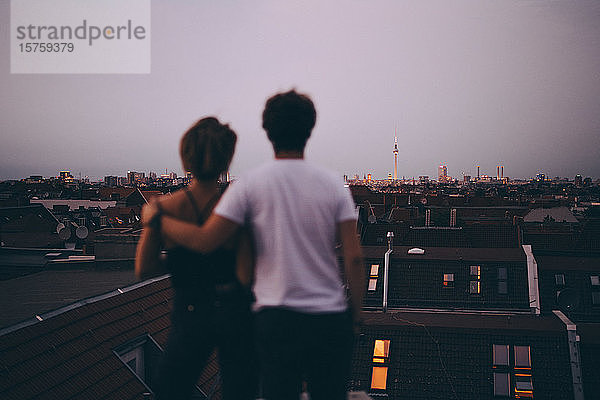 Rückansicht eines Paares  das die Stadt betrachtet  während es in der Dämmerung auf einer Terrasse gegen den Himmel steht