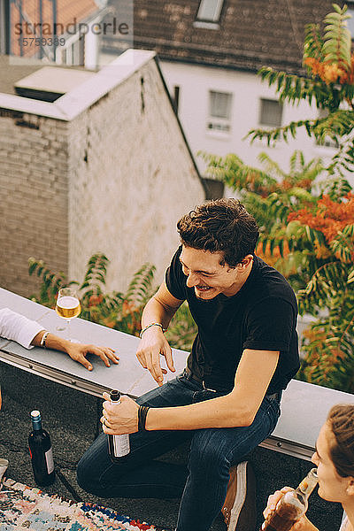 Hochwinkelansicht eines glücklichen Mannes  der Bier genießt  während er bei einer Party mit Freunden auf der Terrasse sitzt