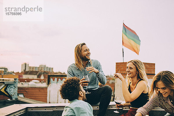 Fröhliche Freunde unterhalten sich beim gemeinsamen Weingenuss auf der Terrasse während einer Party gegen den Himmel