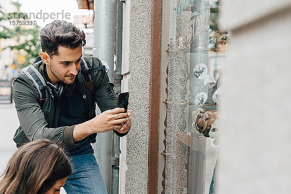 Junger Mann fotografiert Schaufenster mit Mobiltelefon  während er mit einem Freund in der Stadt steht