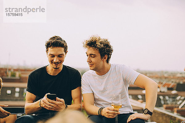 Mann zeigt einem Freund sein Handy und trinkt während der Party auf der Terrasse