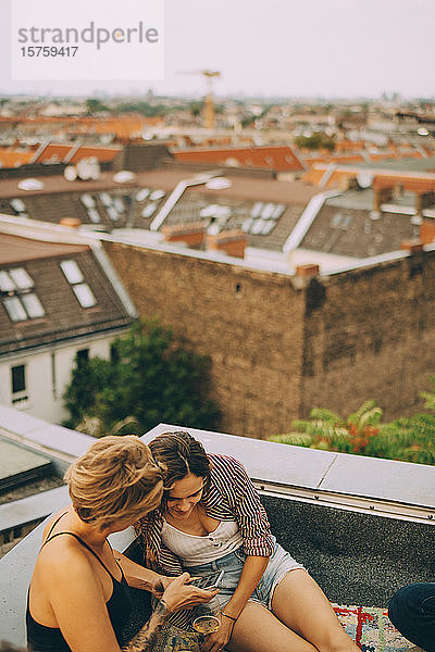 Schrägaufnahme von Freundinnen  die während einer Dachfeier auf einer Terrasse in der Stadt auf ihr Handy schauen