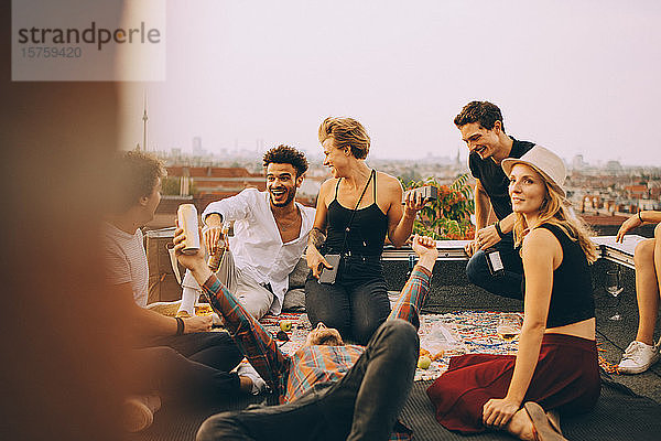 Glückliche Frau spielt Musik über Lautsprecher  während sie mit Freunden auf einer Dachparty genießt