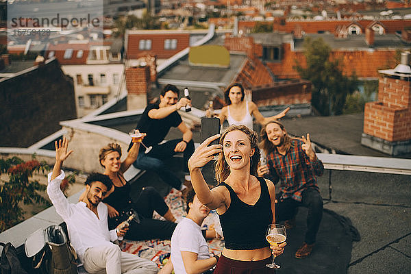 Frau trinkt  während sie sich mit Freunden am Smartphone auf der Terrasse bei einer Party einen Drink genehmigt