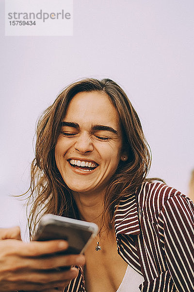 Fröhliche junge Frau betrachtet Smartphone  das von einem Freund auf der Terrasse gehalten wird