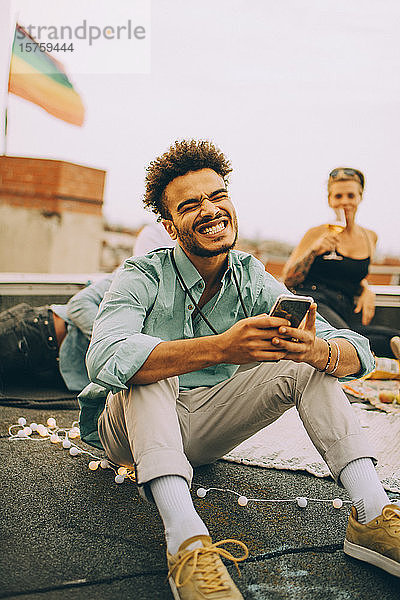 Fröhlicher junger Mann sendet SMS auf Smartphone  während er eine Party auf der Terrasse genießt