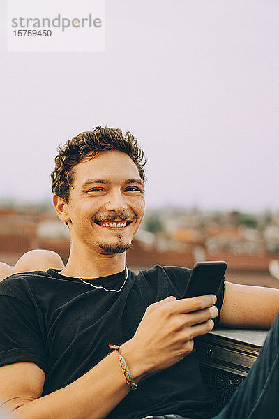 Porträt eines lächelnden jungen Mannes  der ein Mobiltelefon benutzt  während er auf einer Terrasse gegen den Himmel sitzt