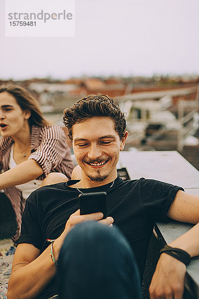 Junger Mann benutzt Smartphone  während ein Freund auf einer Dachparty feiert