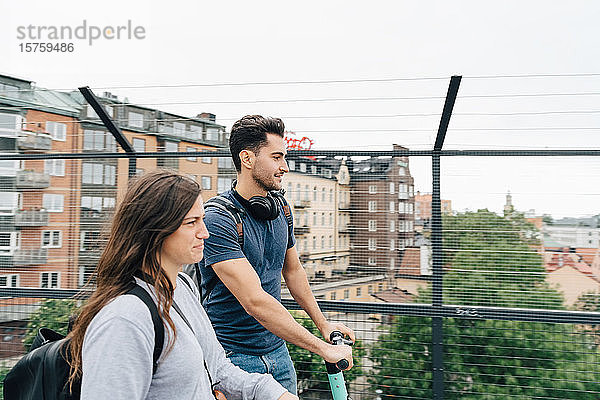 Männliche und weibliche Freunde benutzen Roller auf der Brücke in der Stadt gegen den Himmel