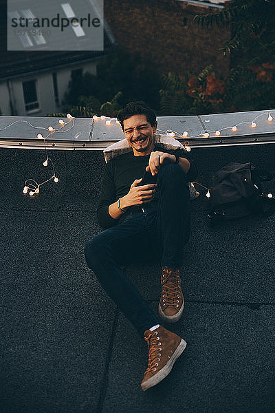 Fröhlicher Mann in voller Länge beim Mobiltelefonieren  während er sich während der Party in der Abenddämmerung auf der beleuchteten Terrasse entspannt