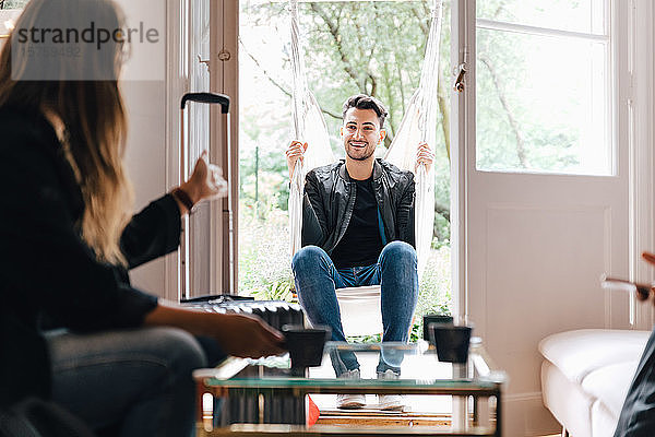 Junge Frau im Gespräch mit einem Freund  der im Wohnzimmer in Schaukel sitzt