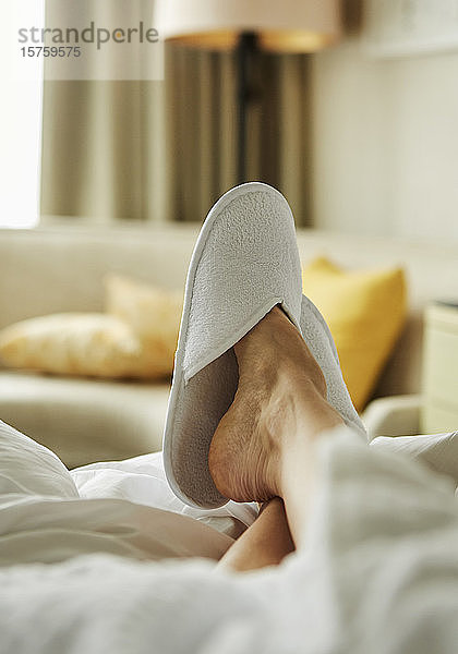 Fuß einer Person mit Schlafzimmerschuh  der im Hotelzimmer auf dem Bett ruht