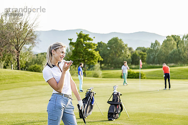Frau spricht mit Smartphone auf dem Golfplatz  Freunde spielen Golf im Hintergrund