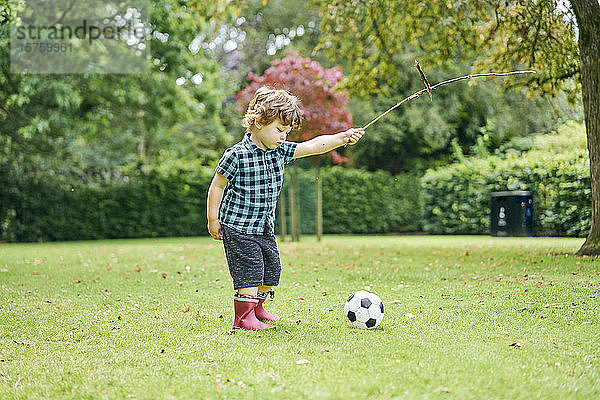 Kleinkind spielt mit Stock und Fussball im Park