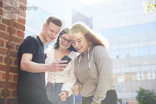 Freunde im Teenageralter benutzen ein Smartphone vor einem sonnigen Schulgebäude