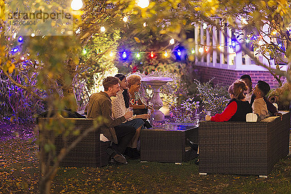 Freunde unterhalten sich und trinken unter Bäumen mit Lichterketten bei einer Gartenparty