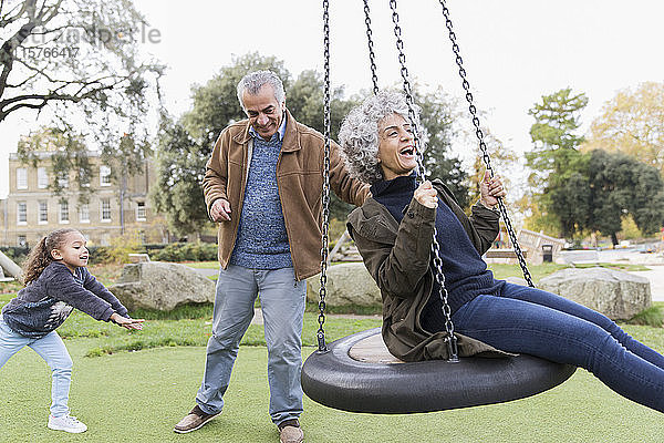 Verspielte Großeltern und Enkelin spielen auf einer Schaukel im Park