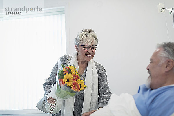 Glückliche ältere Frau bringt Blumenstrauß zu Ehemann erholt sich im Krankenhaus Zimmer