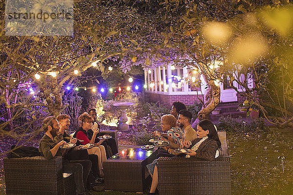 Freunde unterhalten sich und essen Nachtisch unter Bäumen mit Lichterketten bei einer Gartenparty