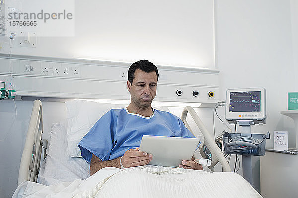 Männlicher Patient  der ein digitales Tablet benutzt und sich im Krankenhausbett ausruht