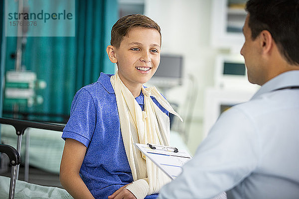 Männlicher Kinderarzt im Gespräch mit einem jungen Patienten mit Arm in einer Schlinge in einer Klinik