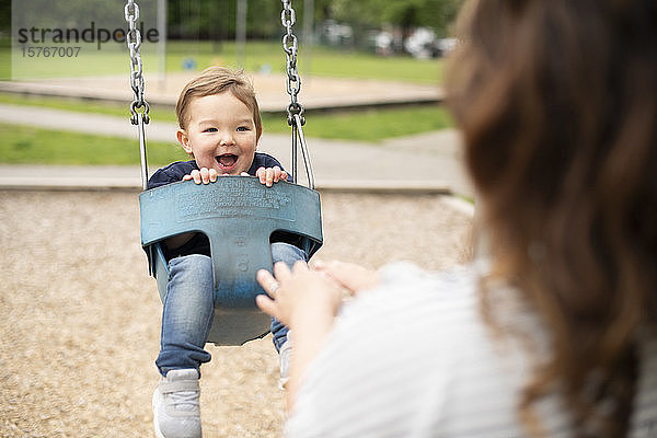 Mutter schiebt glückliche niedliche Kleinkind Mädchen in Schaukel auf dem Spielplatz