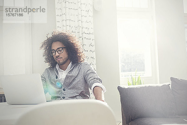 Junger Mann arbeitet am Laptop im sonnigen Wohnzimmer