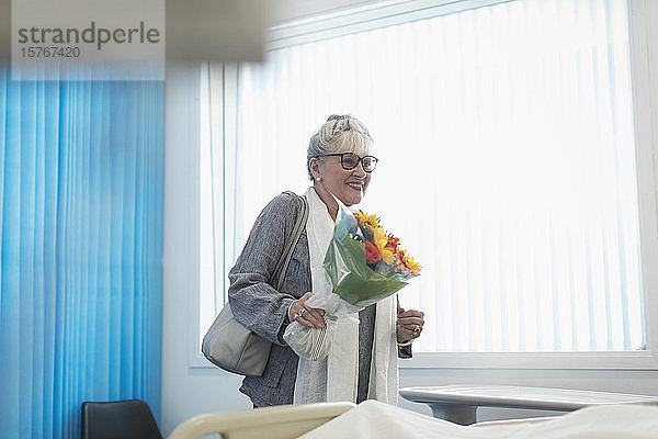 Lächelnde ältere Besucherin mit Blumenstrauß im Krankenhauszimmer