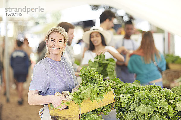 Porträt einer lächelnden  selbstbewussten Frau  die auf einem Bauernmarkt eine Gemüsekiste trägt