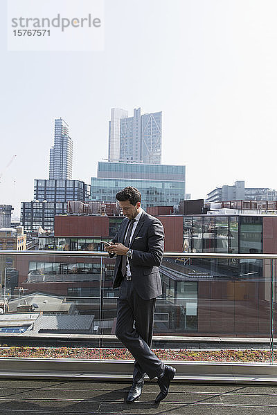 Geschäftsmann  der auf einem sonnigen  städtischen Hochhausbalkon ein Smartphone benutzt