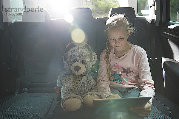 Mädchen mit Teddybär benutzt digitales Tablet auf dem Rücksitz eines Autos