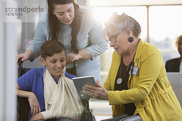 Arzt mit digitalem Tablet im Gespräch mit einem jungen Patienten im Rollstuhl und seiner Mutter in der Kliniklobby