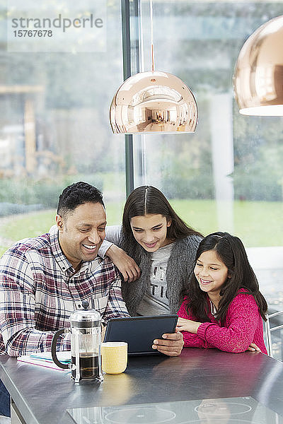 Ein glücklicher Vater und seine Töchter benutzen ein digitales Tablet in der morgendlichen Küche