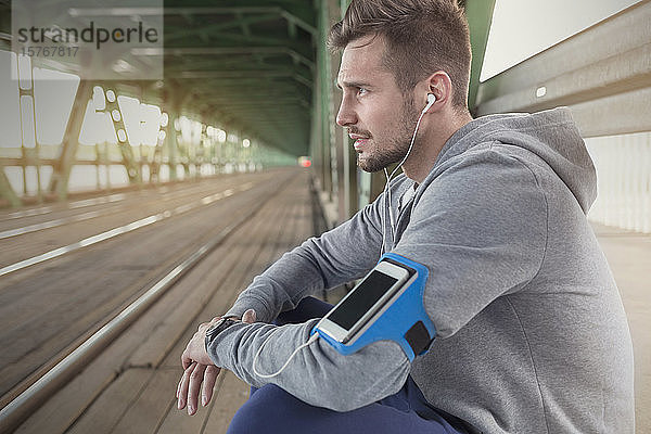 Junger männlicher Läufer  der sich ausruht und mit Kopfhörern und mp3-Player auf dem Bahnsteig Musik hört