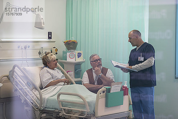 Arzt mit Krankenblatt im Gespräch mit einem älteren Ehepaar im Krankenhauszimmer
