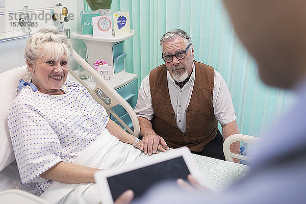 Arzt mit digitalem Tablet im Gespräch mit einem älteren Ehepaar im Krankenhauszimmer
