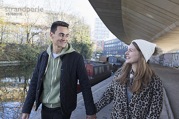 Glückliches junges Paar  das sich an den Händen hält und an einem städtischen Kanal entlanggeht