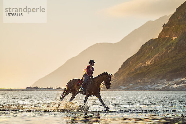 Junge Frau im Galopp auf einem Pferd in der ruhigen Meeresbrandung