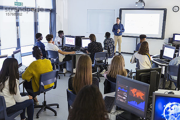 Schüler der Mittelstufe an Computern  die den Lehrer auf der Projektionsfläche im Klassenzimmer beobachten
